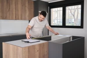 Man measuring kitchen cabinetry with a tape measure, preparing for installation in a modern kitchen setting.